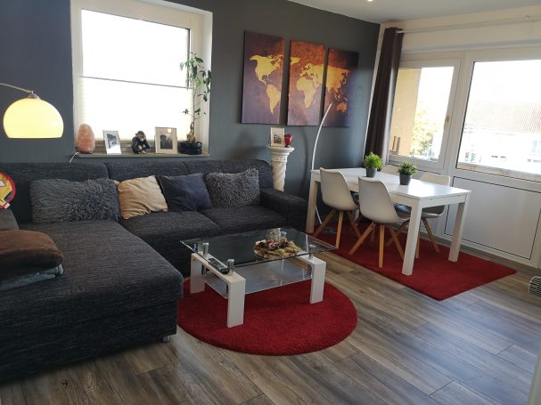 Wohnzimmer mit grauem Sofa, Tisch, Pflanzen und bunten Wandbildern. Rote Teppiche.