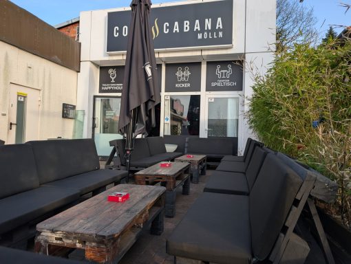 Gemütliche Lounge mit schwarzen Sitzbänken und Holztischen vor einem Restaurant.