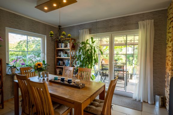 Esszimmer mit Holztisch, sechs Stühlen, Regal, großen Fenstern und Blick in den Garten