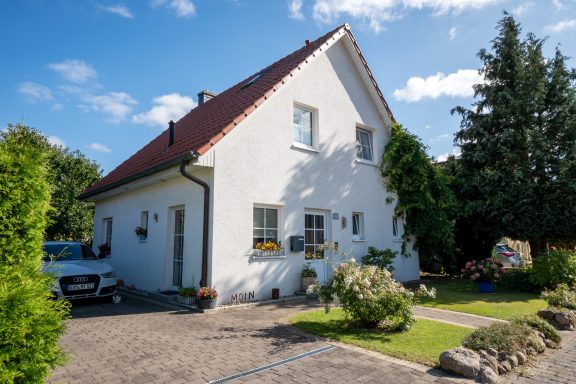 Weißes Einfamilienhaus mit rotem Satteldach, Vorgarten, Einfahrt und blauem Himmel