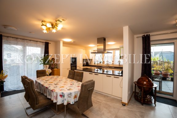 Moderne Küche mit Esstisch und Stühlen, helle Fenster mit Pflanzen im Hintergrund.