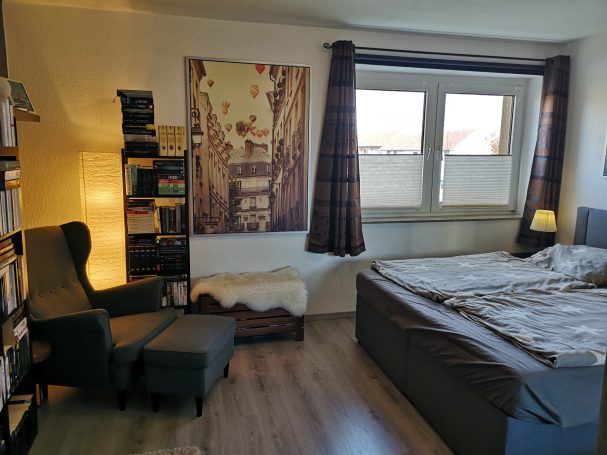 Gemütliches Schlafzimmer mit einem Bett, Bücherregal, Sessel und großem Fenster.