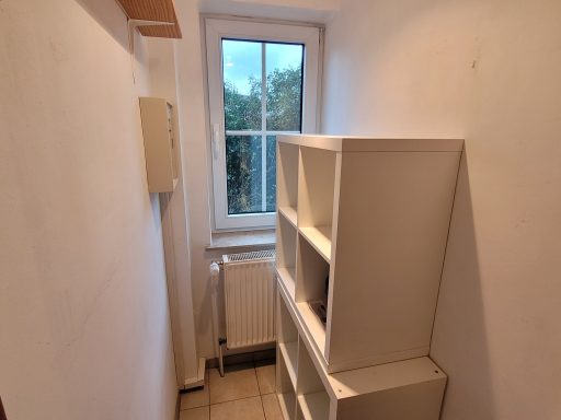 Ein heller Raum mit einem Fenster und einem weißen Bücherregal in der Ecke.