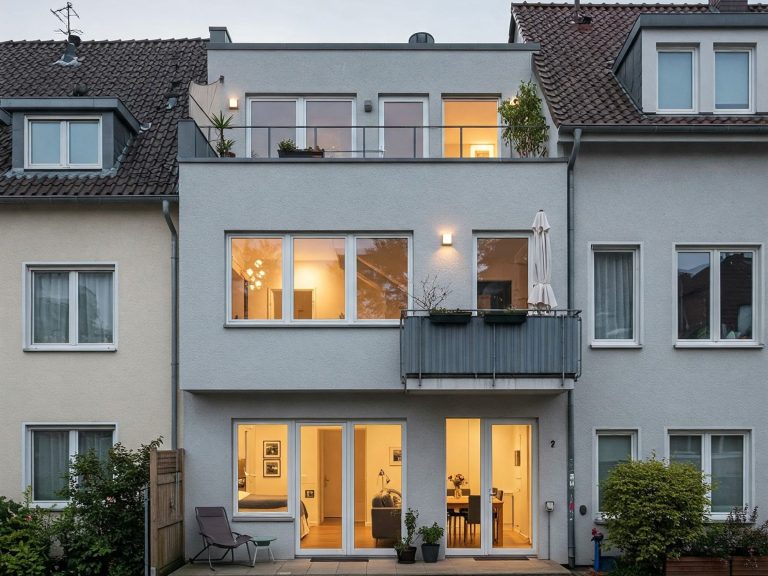 Moderne Wohnhausfassade mit großen Fenstern und Balkon, warm beleuchtet.