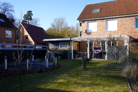 Blick auf ein Haus mit Garten, umgeben von Bäumen und einem klaren blauen Himmel.