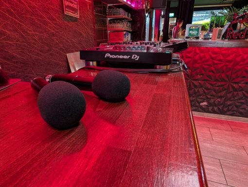Zwei schwarze Mikrofone liegen auf einem Holztisch vor einer DJ-Konsole in einem farbigen Raum.