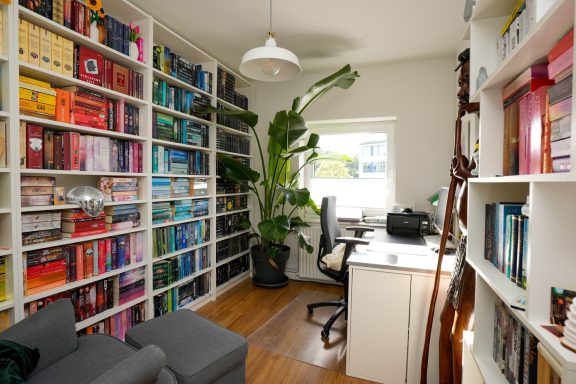 Büro mit Regalen voller Bücher, Schreibtisch und Pflanzen in einem hellen Raum.