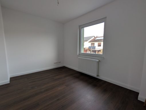 Helles, leeres Zimmer mit einer Fensteransicht auf ein Wohngebiet. Holzfußboden.