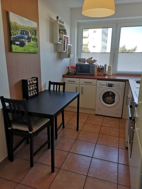Kleine Küche mit Tisch, Stühlen, Waschmaschine und Fenster. 